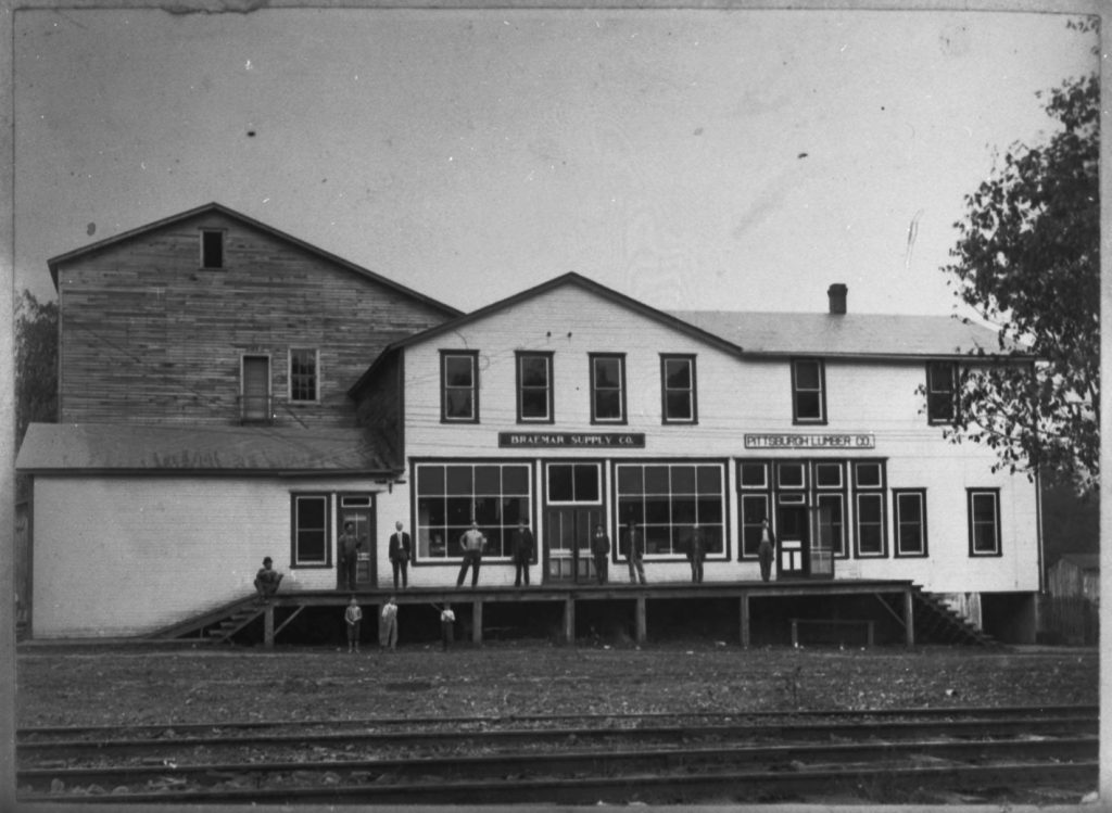 Εξωτερική όψη της εταιρείας Lumber Company Pittsburgh και Braemar Supply Company Building το 1915. Οι άνδρες και τα αγόρια στέκονται έξω από το κτίριο. Οι διαδρομές σιδηροδρόμων εμφανίζονται στο προσκήνιο.